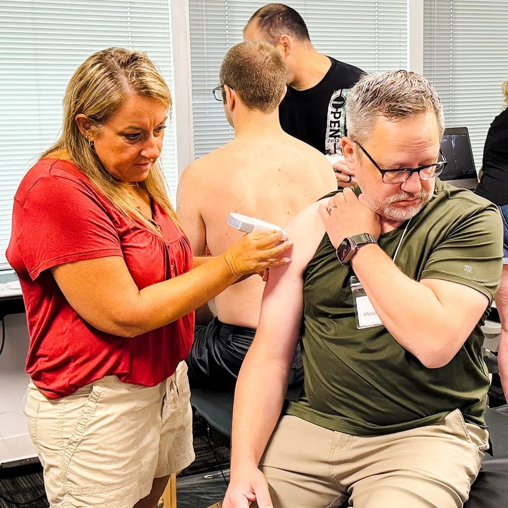 Instructor guiding an ultrasound probe over a live model’s shoulder during an injection training class